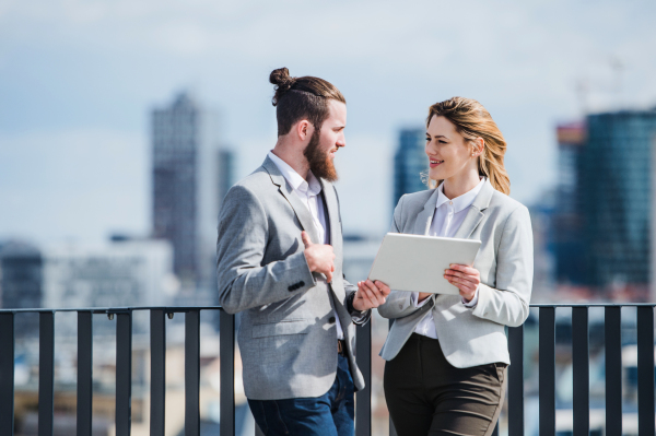 Two cheerful young business people with tablet standing on a terrace outside office, working.