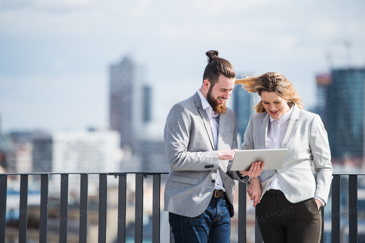 Two cheerful young business people with tablet standing on a terrace outside office, working.