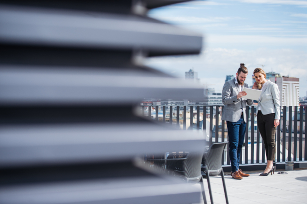 Two cheerful young business people with tablet standing on a terrace outside office, working.