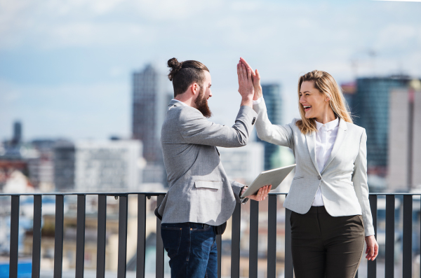 Two young business people with tablet standing on a terrace outside office, expressing excitement.