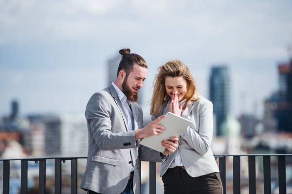 Two cheerful young business people with tablet standing on a terrace outside office, working.