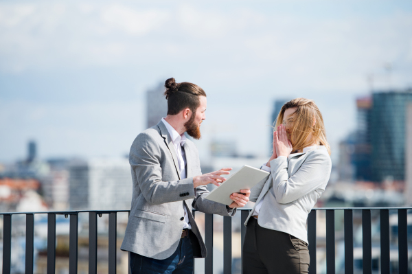 Two cheerful young business people with tablet standing on a terrace outside office, working.