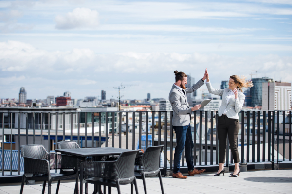 Two young business people with tablet standing on a terrace outside office, expressing excitement.