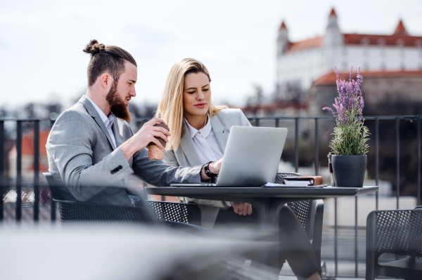 Two cheerful young business people with laptop sitting on a terrace outside office, working.