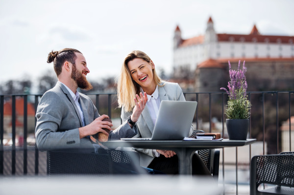 Two cheerful young business people with laptop sitting on a terrace outside office, working.