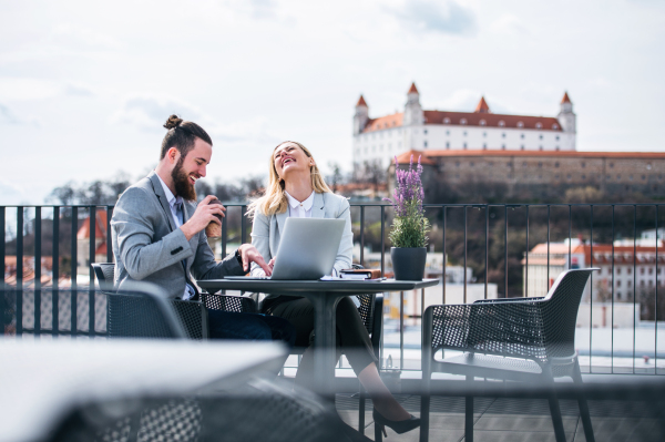 Two cheerful young business people with laptop sitting on a terrace outside office, laughing when working.