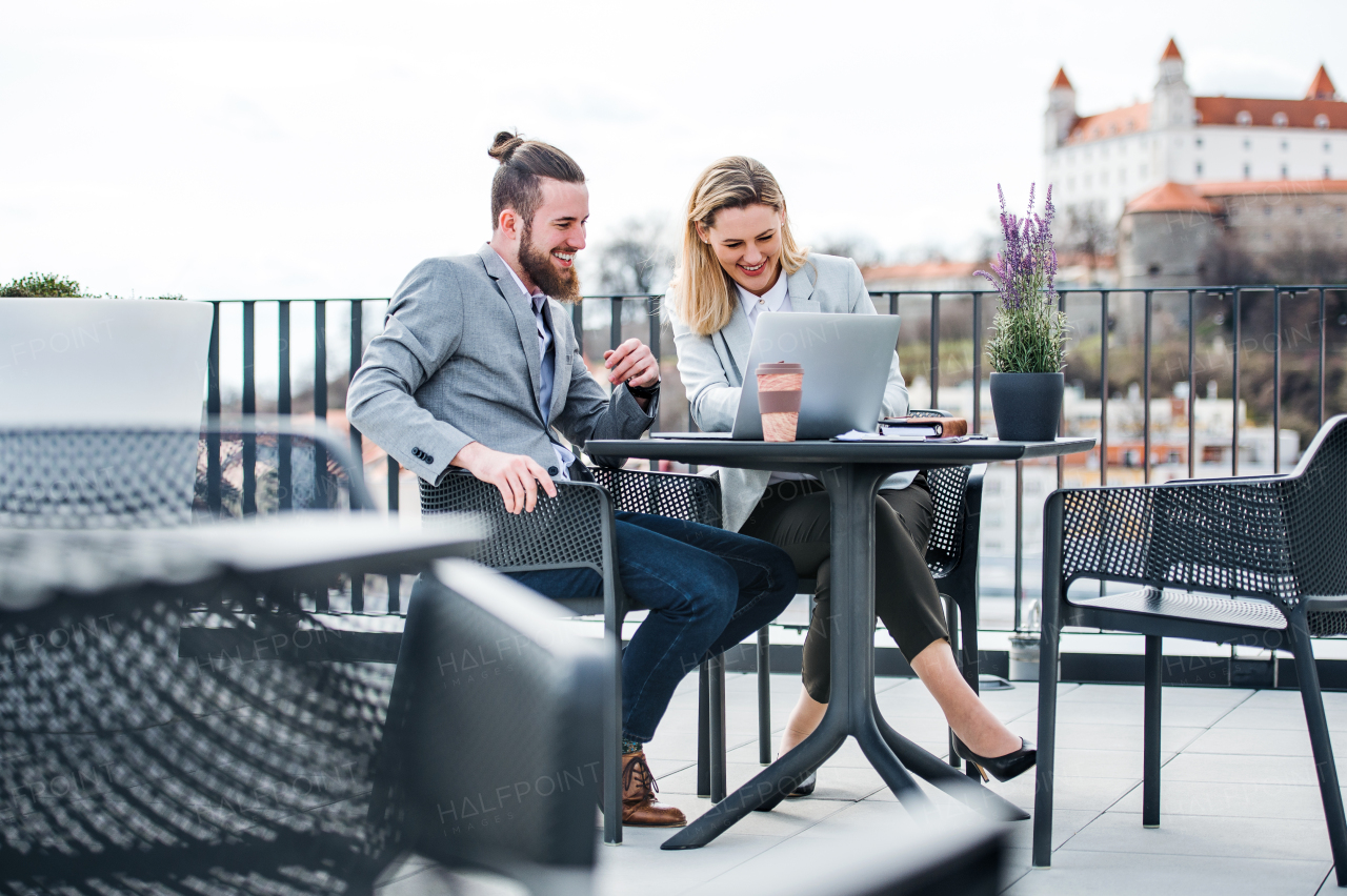 Two cheerful young business people with laptop sitting on a terrace outside office in Bratislava, working.