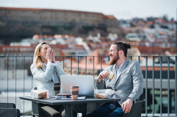 Two young business people sitting at the table on a terrace outside office, having lunch break.