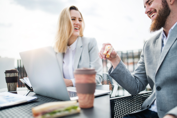 Two cheerful young business people with laptop sitting on a terrace outside office, working and eating snack.