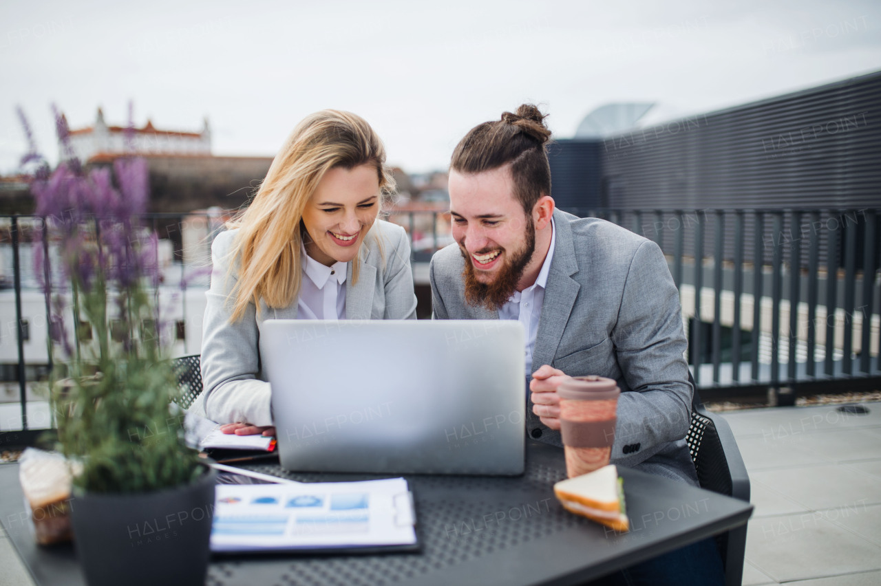 Two cheerful young business people with laptop sitting on a terrace outside office, working.