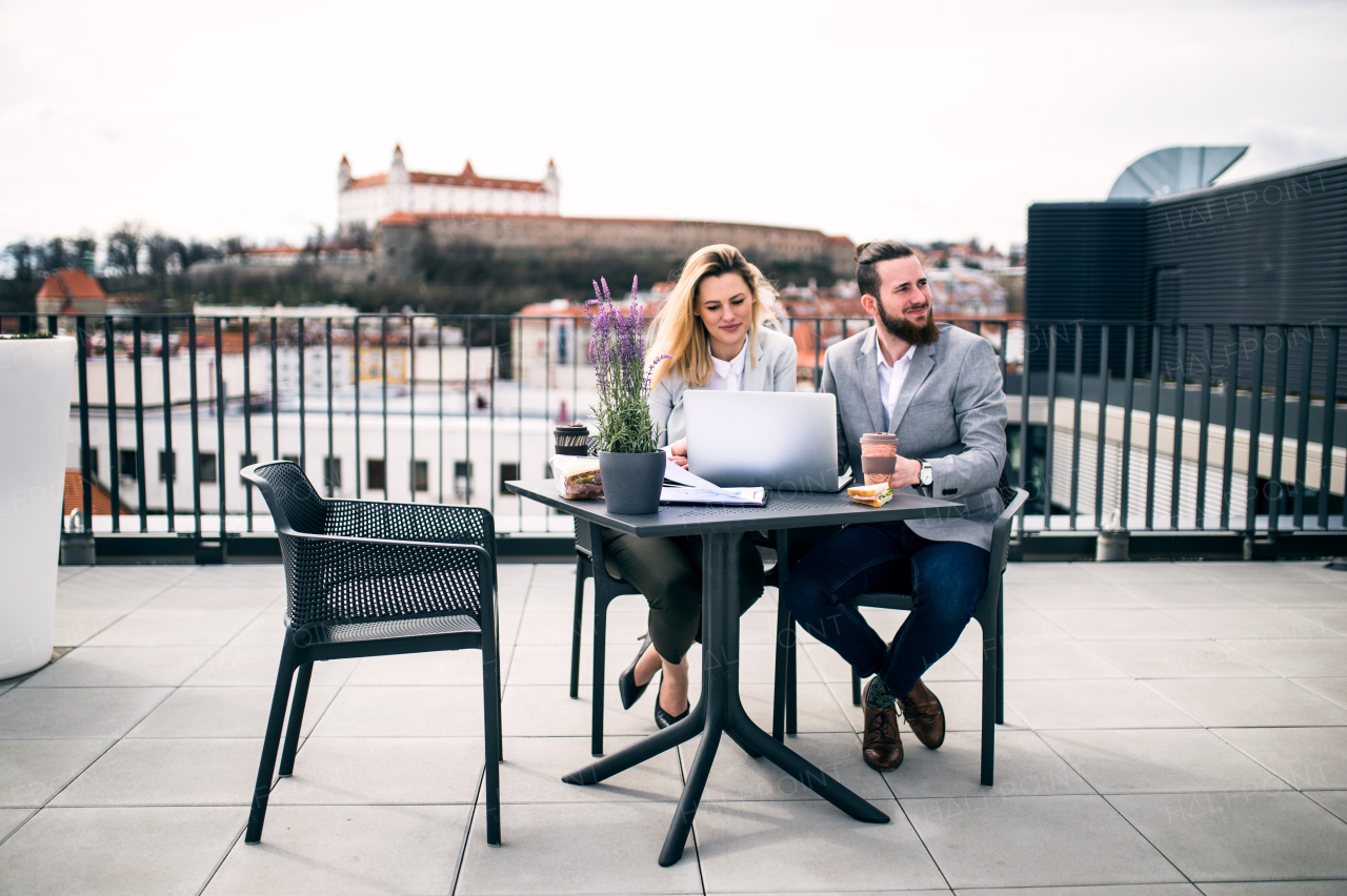 Two cheerful young business people with laptop sitting on a terrace outside office, working.