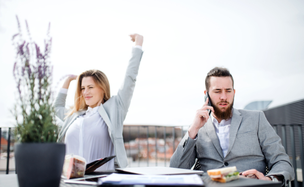 Two cheerful young business people with laptop sitting on a terrace outside office, working.