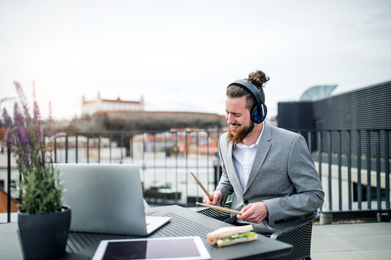 A young businessman with headphones and drumsticks sitting on a terrace, having fun.