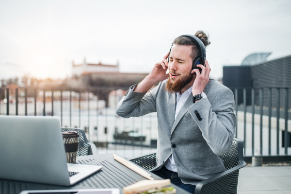 A young businessman with laptop, headphones and snack sitting on a terrace, having break.