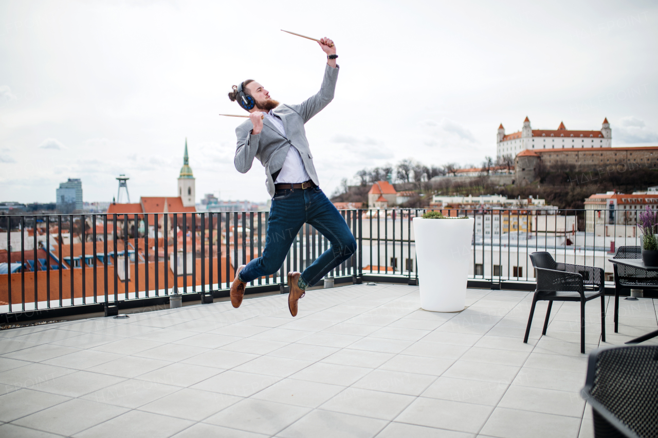 A young businessman with headphones and drumsticks standing on a terrace, having fun.