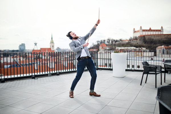 A young businessman with headphones and drumsticks standing on a terrace, having fun.