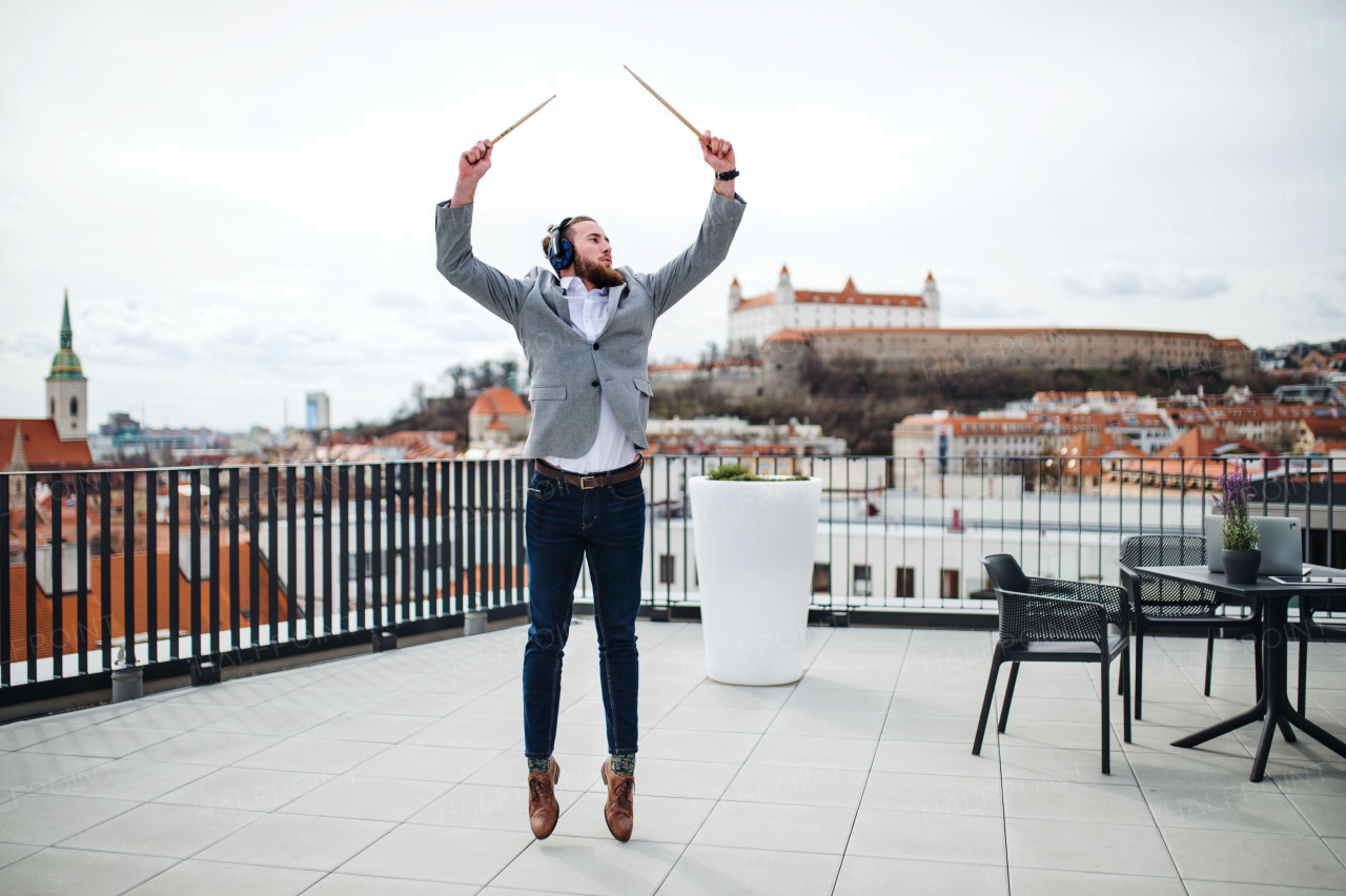 A young businessman with headphones and drumsticks standing on a terrace, having fun.