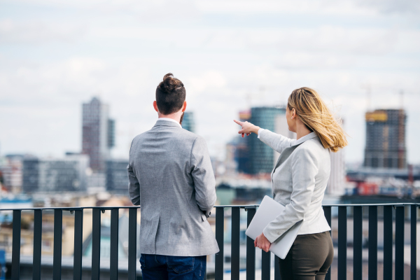 Rear view of two cheerful young business people with laptop standing on a terrace outside office, talking.