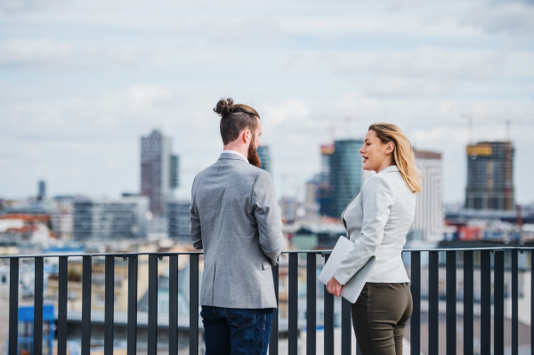 Two cheerful young business people with tablet standing on a terrace outside office, talking.