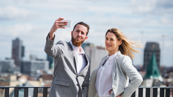 Two cheerful young business people with smartphone standing on a terrace outside office, taking selfie.