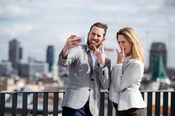 Two cheerful young business people with smartphone standing on a terrace outside office, taking selfie.