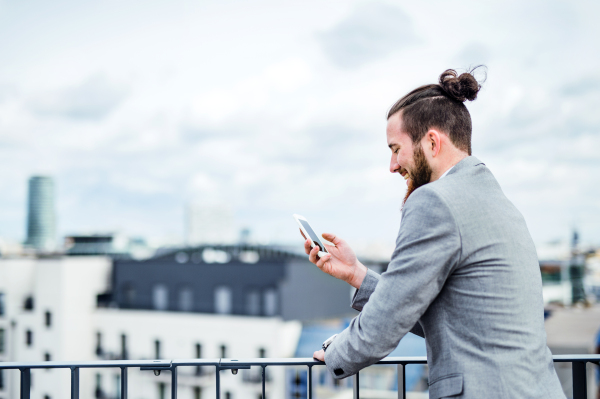 A young businessman with smartphone standing on a terrace outside office, text messaging. Copy space.