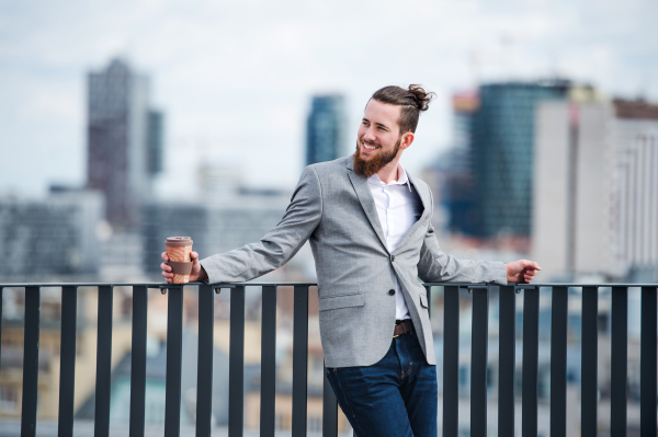 A young businessman with coffee standing on a terrace outside office, resting.