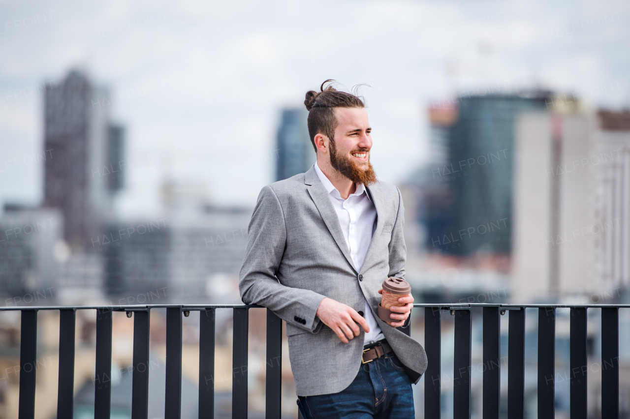 A young businessman with coffee standing on a terrace outside office, resting.