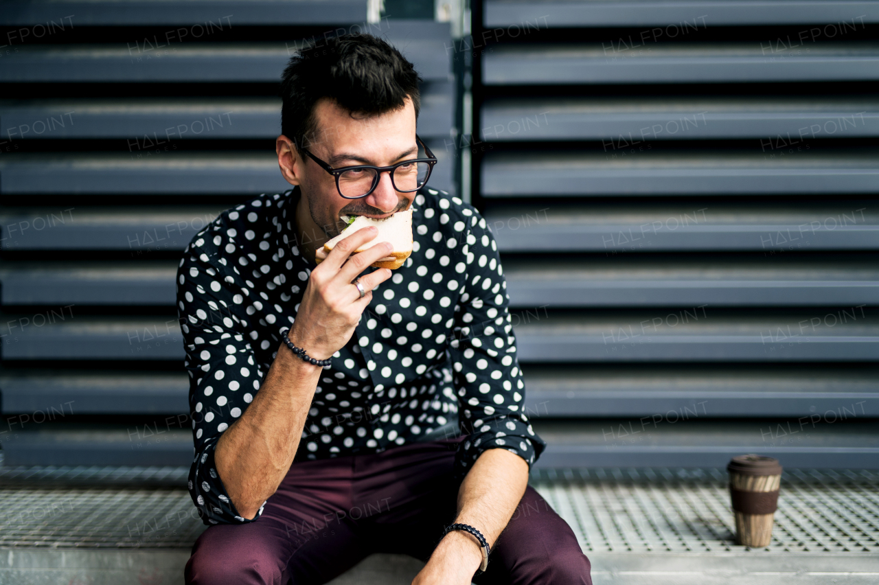 A young businessman with coffee and sandwich sitting outdoors, having a snack and resting.