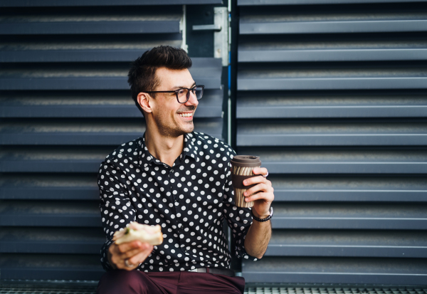 A young businessman sitting outdoors, drinking coffee and eating sandwich. Copy space.