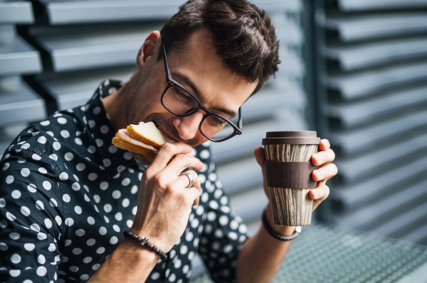 A young businessman sitting outdoors, drinking coffee and eating sandwich.