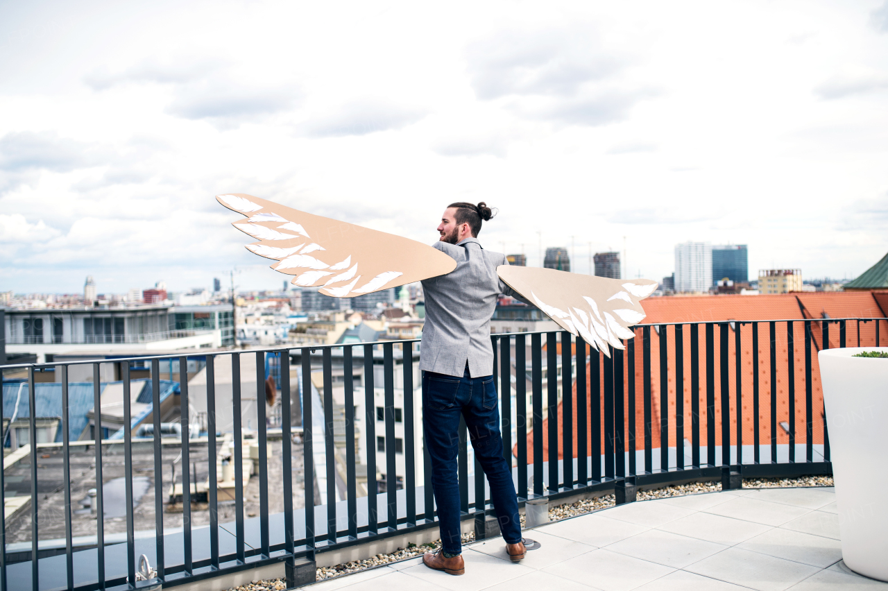 A rear view of young businessman with cardboard wings standing on a terrace, flying metaphor and freedom concept.