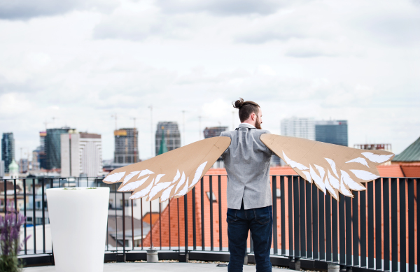 A young businessman with cardboard wings standing on a terrace, flying metaphor and freedom concept. A rear view.