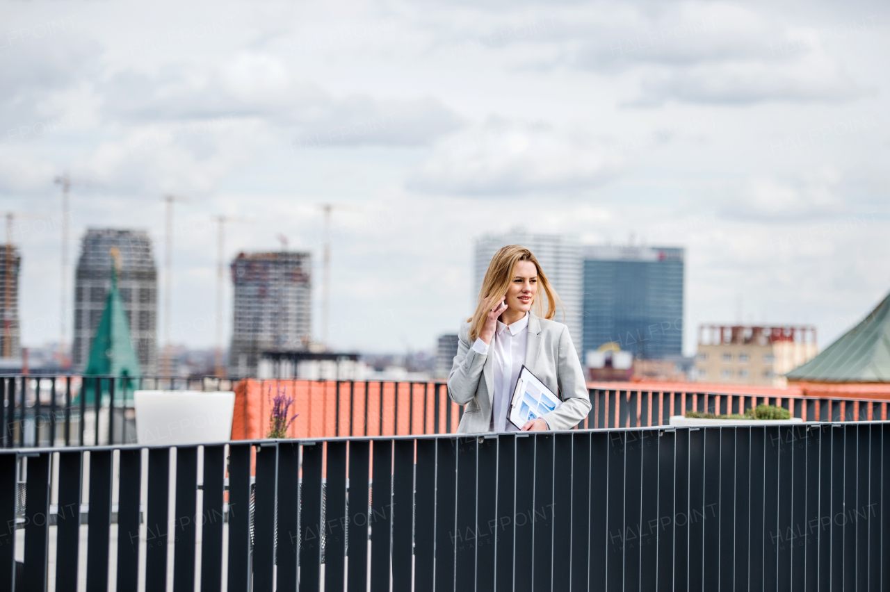 A young businesswoman with smartphone and laptop standing on a terrace, making a phone call when working.