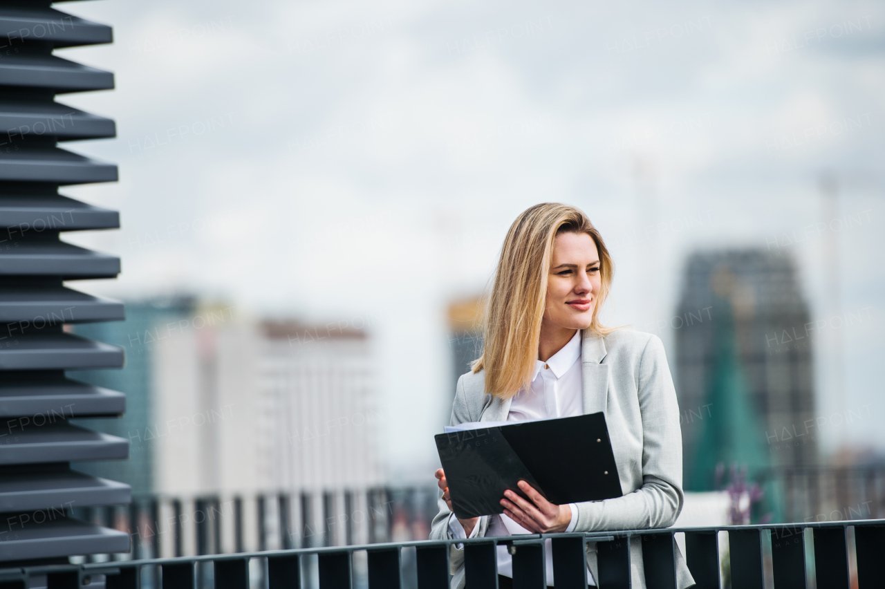 A portrait of young blond businesswoman with clipboard standing outdoors on terrace outside office. Copy space.