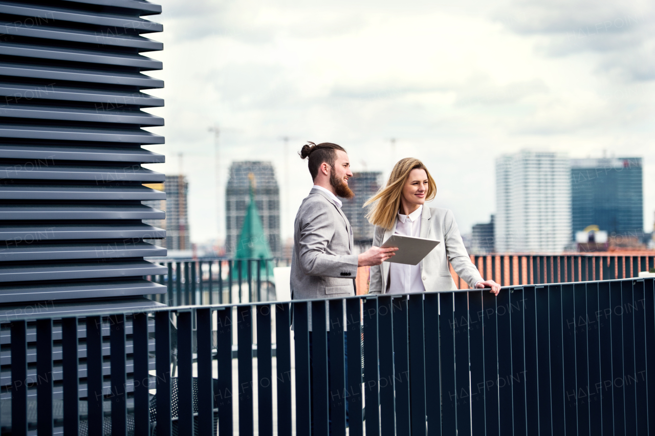 Two cheerful young business people with tablet standing on a terrace outside office, working.