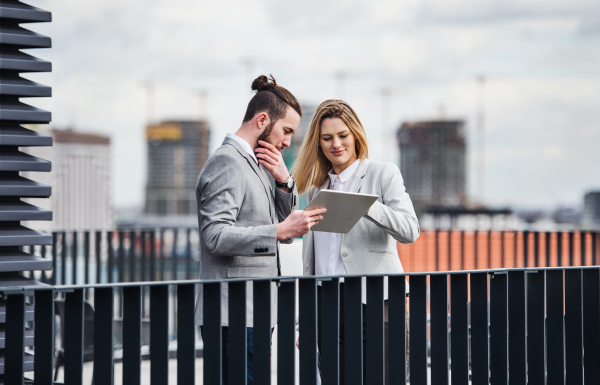 Two cheerful young business people with tablet standing on a terrace outside office, working.