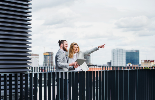 Two cheerful young business people with tablet standing on a terrace outside office, working.