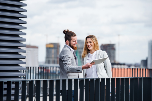 Two cheerful young business people with tablet standing on a terrace outside office, working.