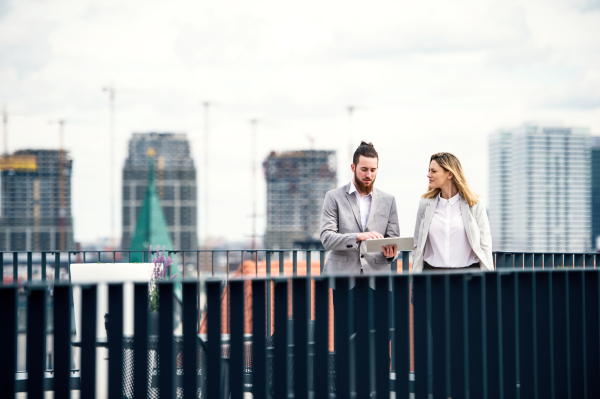 Two cheerful young business people with tablet standing on a terrace outside office, working.