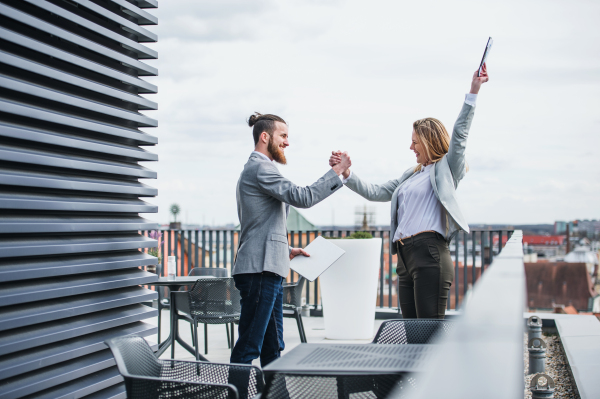 Two young business people with tablet standing on a terrace outside office, expressing excitement.