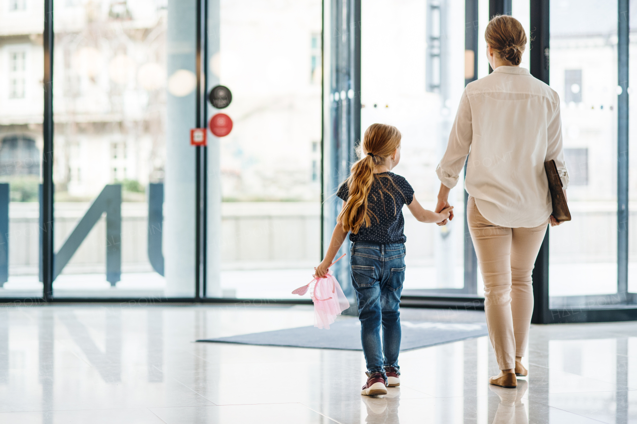 A rear view of businesswoman with small daughter walking in office building, holding hands.