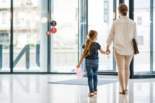 A rear view of businesswoman with small daughter walking in office building, holding hands.