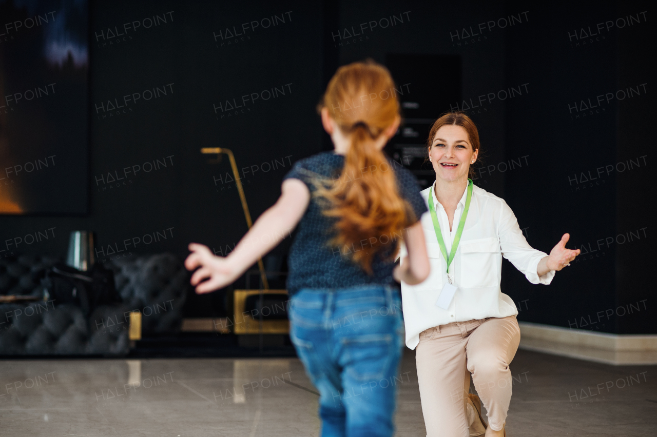 A businesswoman greeting with happy small daughter after a day in nursery in office building.