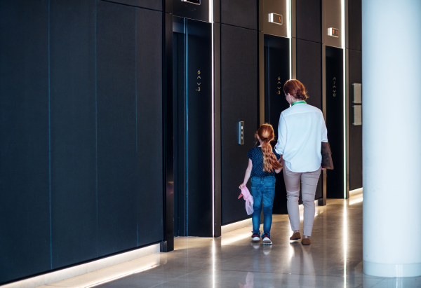 A side view of businesswoman with small daughter walking by elevators in office building.