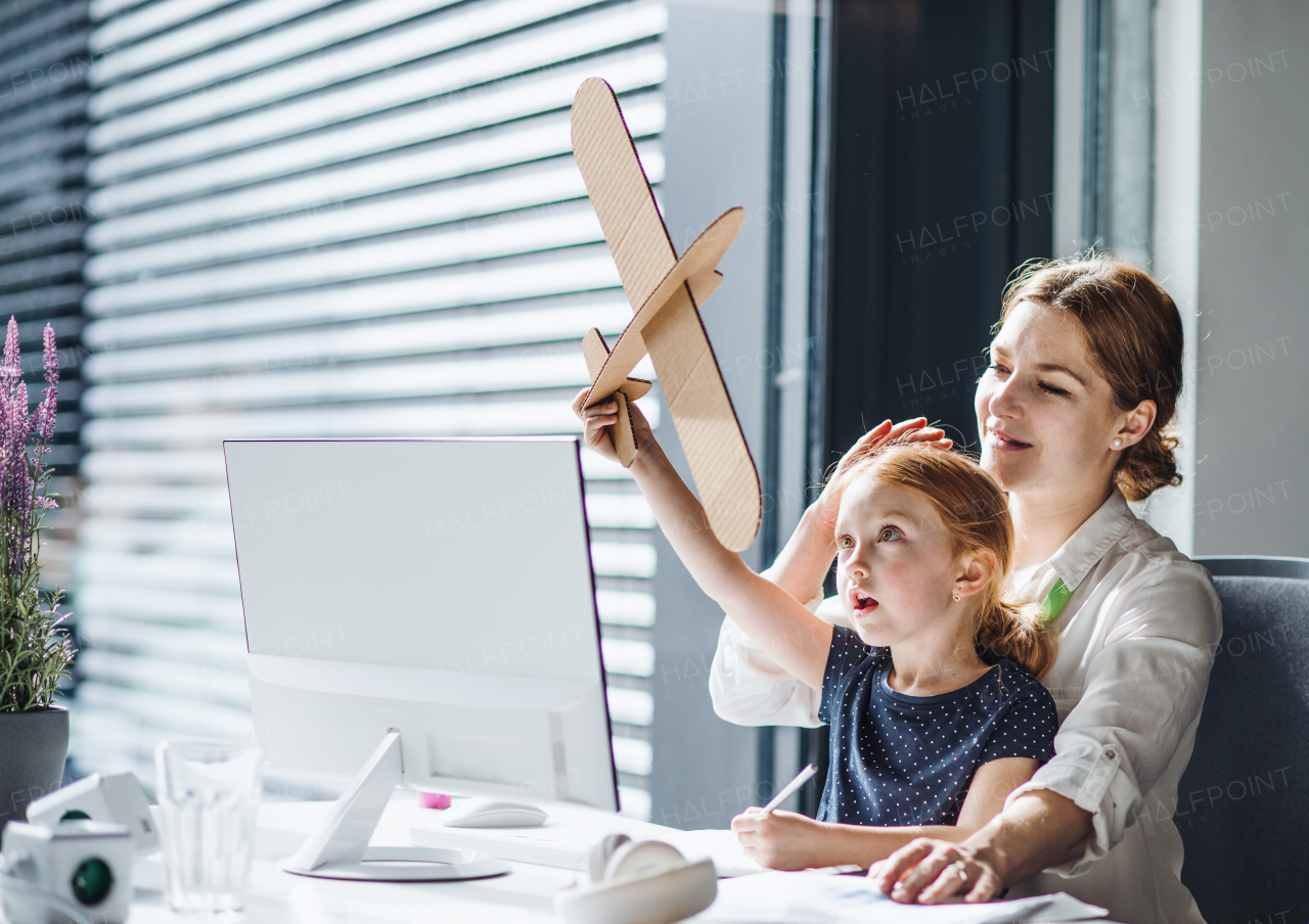 A businesswoman sitting and working in an office, a small daughter playing with a paper plane.