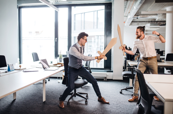 Two young businessmen with cardboard swords in an office, having fun. A competition concept.