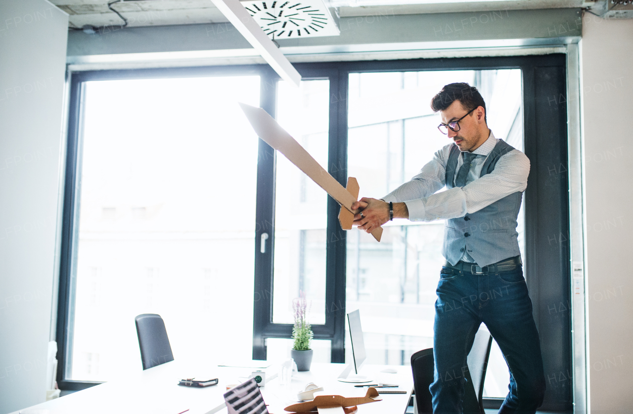 A young businessman with cardboard sword in an office, having fun. A competition concept.