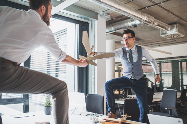 Two young businessmen with cardboard swords in an office, having fun. A competition concept.