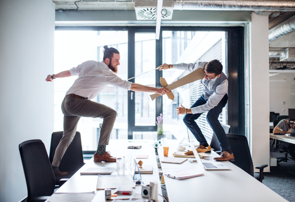 Two young businessmen with cardboard swords in an office, having fun. A competition concept.